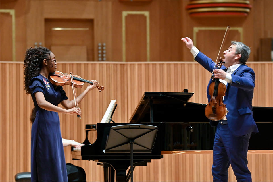 Man wearing a blue suit, holding a violin, with a female student playing a violin, with a piano behind them.